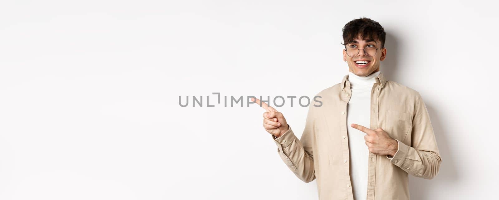 Excited and enthusiastic young man in glasses, pointing fingers and looking left with happy smile, standing in awe on white background.