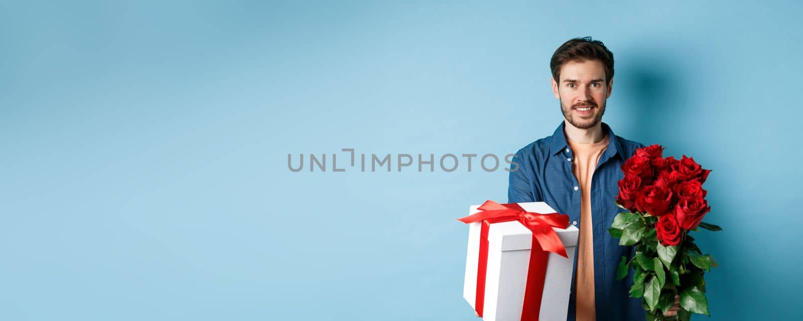 Love and Valentines day concept. Charming young man giving gift and bouquet of roses to girlfriend, standing over blue background by Benzoix