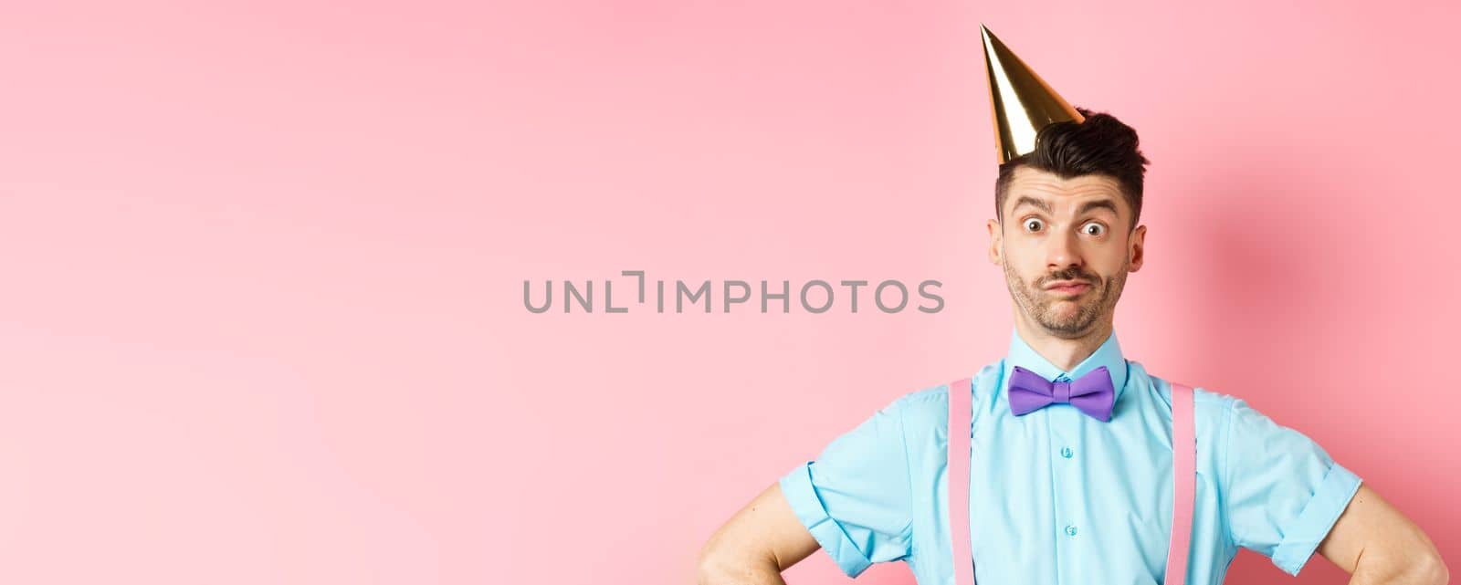 Holidays and celebration concept. Close-up of confused male entertainer in party hat and bow-tie, looking puzzled and shocked, standing over pink background.