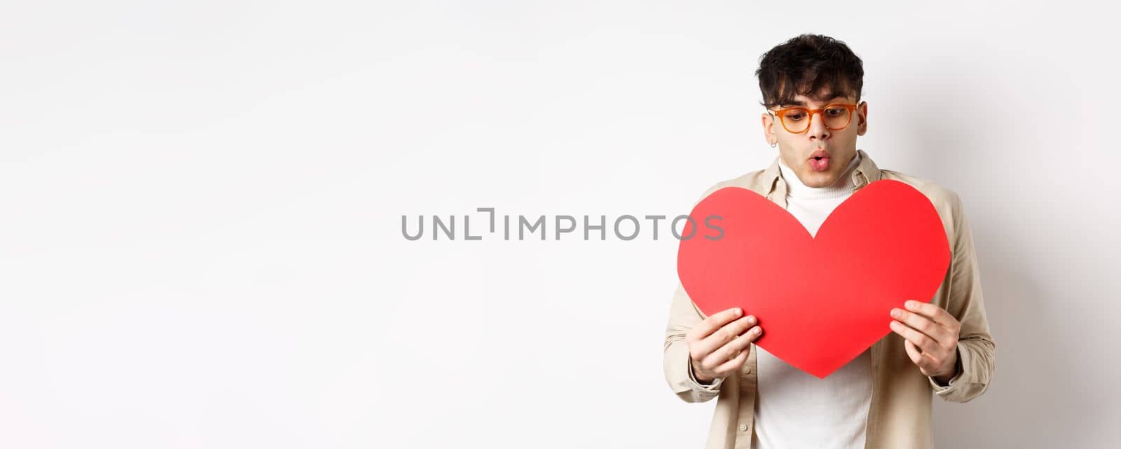 Surprised handsome man receive big red heart postcard on Valentines day, looking at gift with amazement, enjoying lovers day, standing over white background by Benzoix