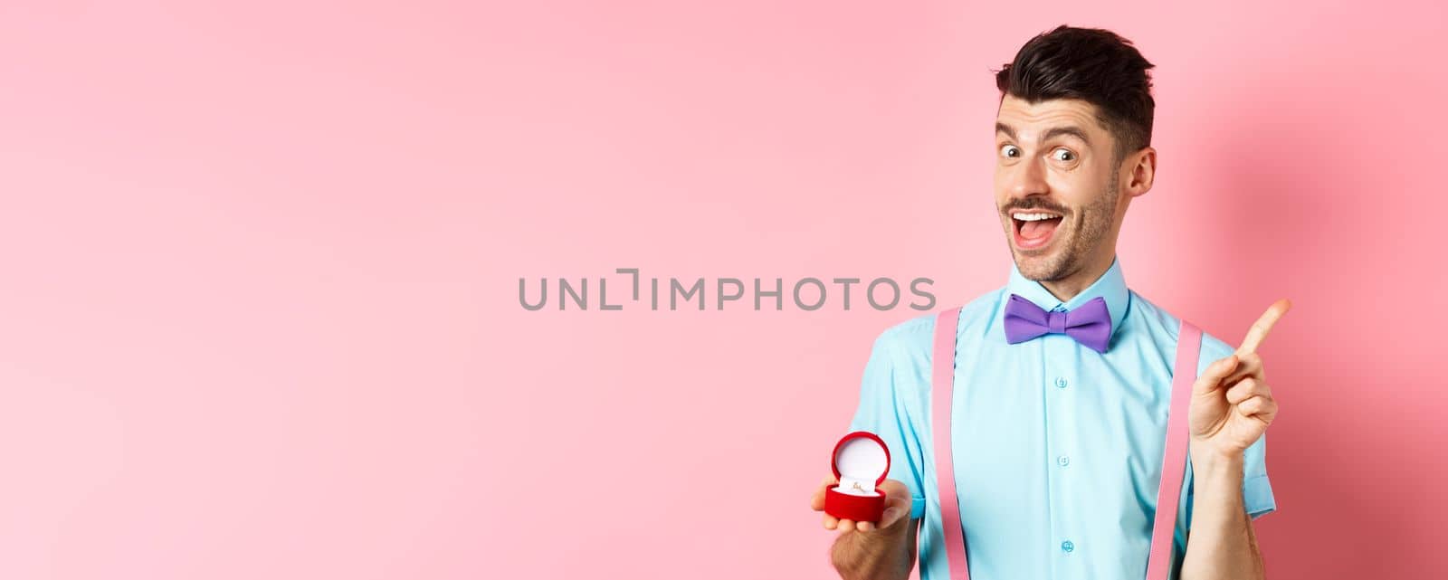 Valentines day. Funny guy with moustache and bow-tie, showing engagement ring and pointing finger at upper right corner, standing on pink background.