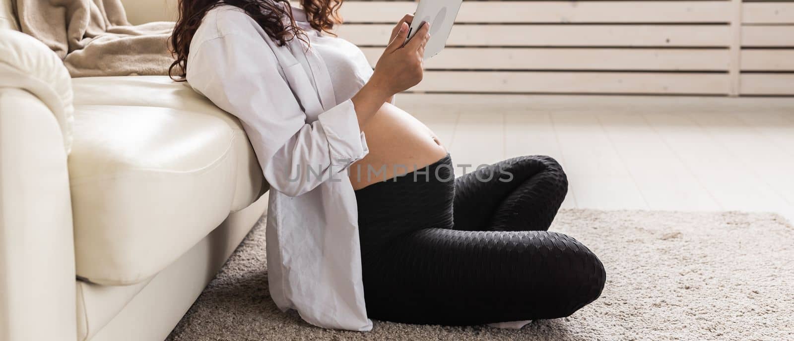 Pregnant woman holding tablet sitting on a carpet near a couch in the living room at home.