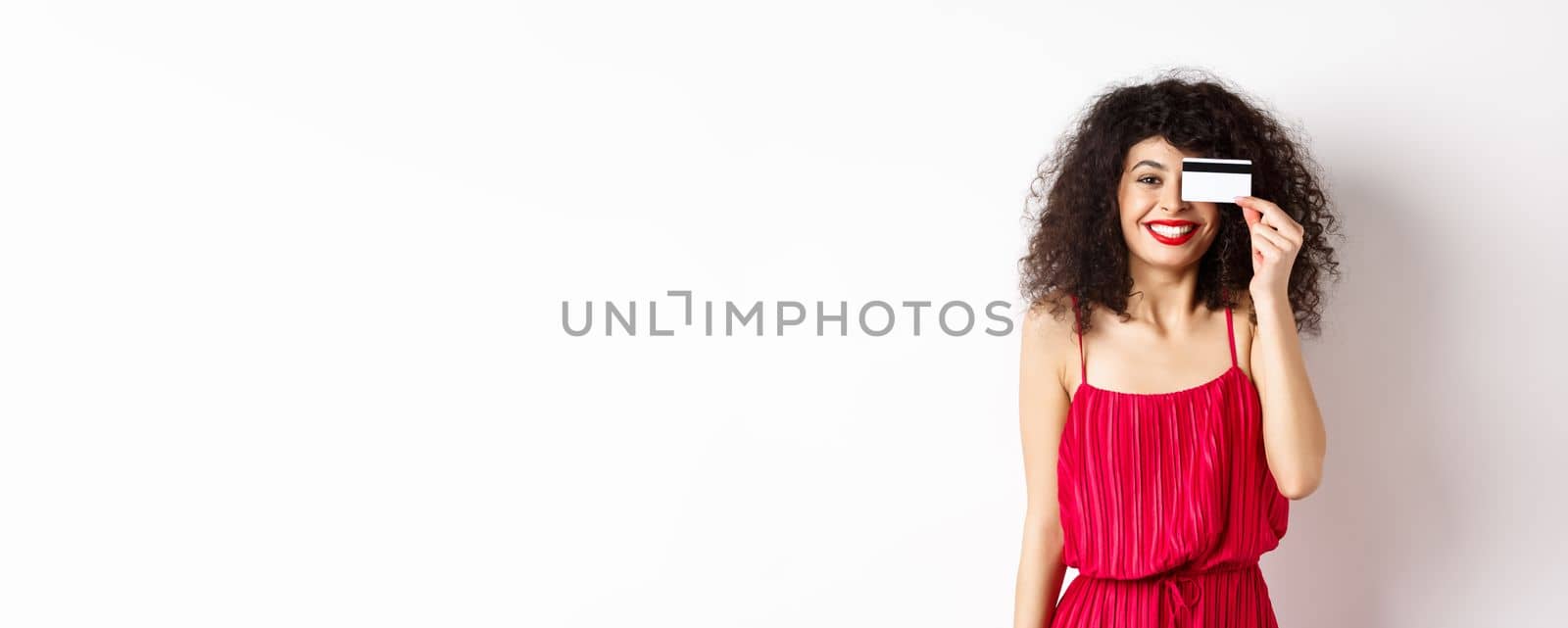 Image of elegant woman in red dress, showing plastic credit card and smiling at camera, standing over white background by Benzoix