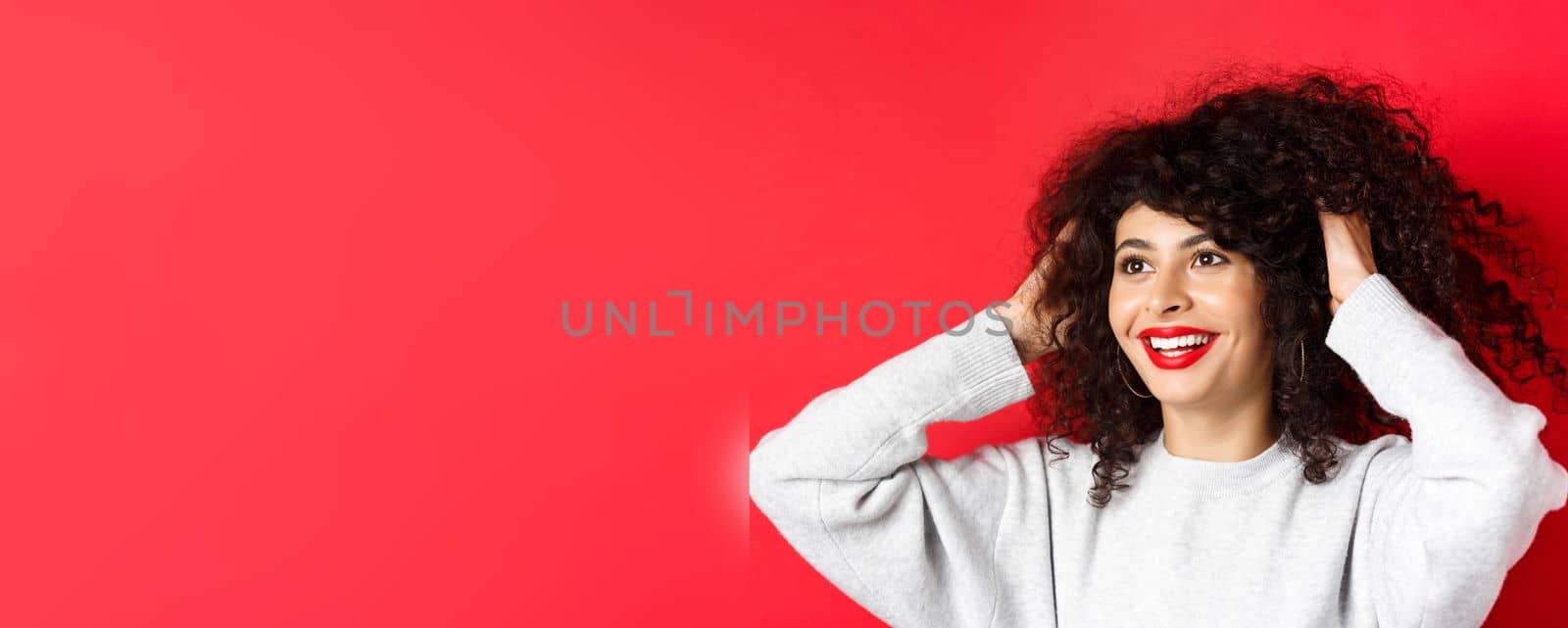 Happy beautiful woman touching natural curly hair and smiling pleased, looking left, like her hairstyle, standing against red background.