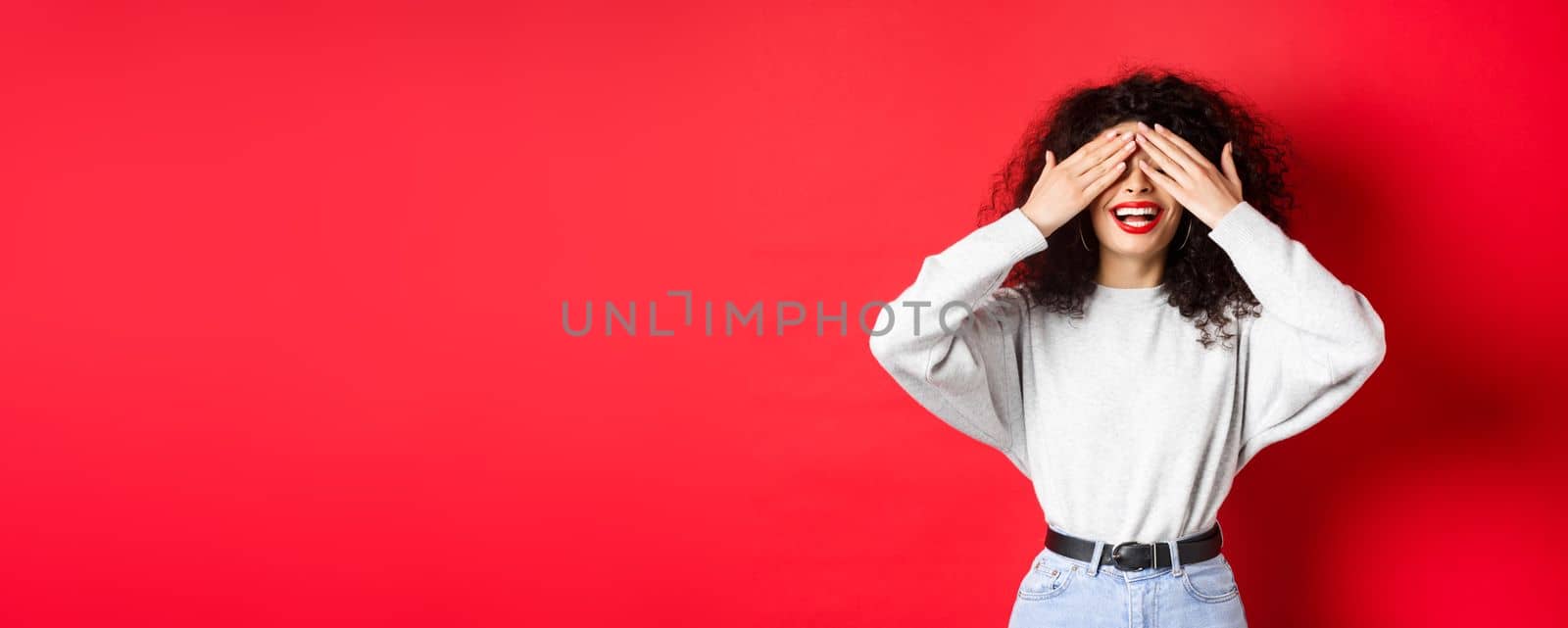 Happy beautiful woman with curly hair and red lips, covering eyes with hands and waiting surprise, smiling excited, standing against red background by Benzoix