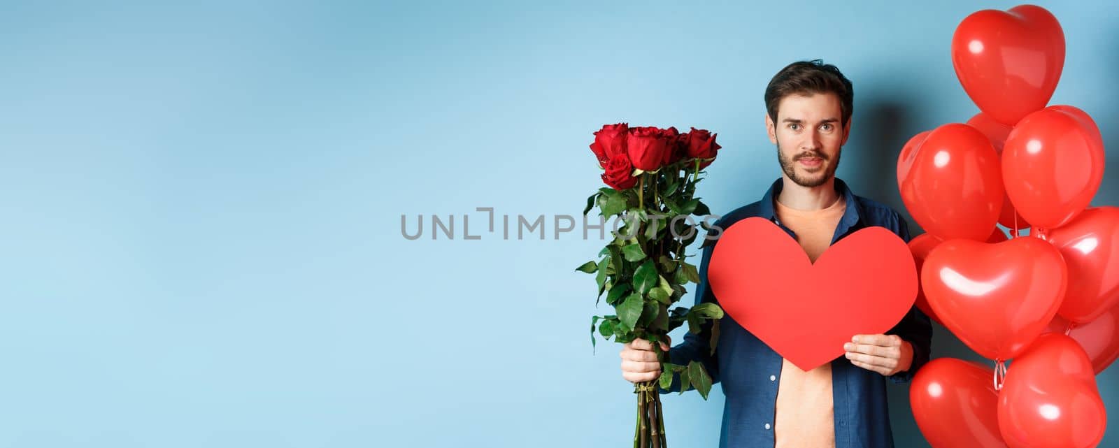 Valentines day romance. Young man with bouquet of red roses and heart balloons smiling, bring presents for lover on valentine date, standing over blue background by Benzoix