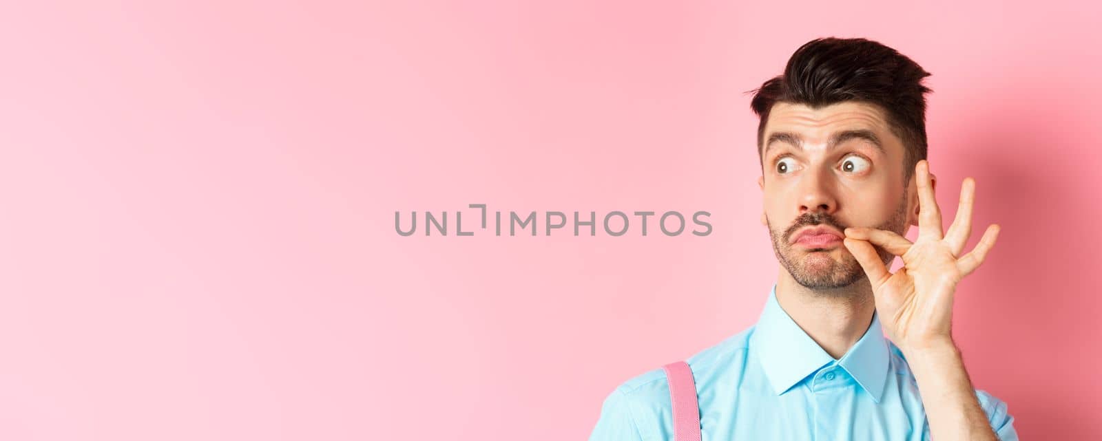 Close up of young caucasian man touching moustache and pucker lips, staring left at logo, standing on pink background.