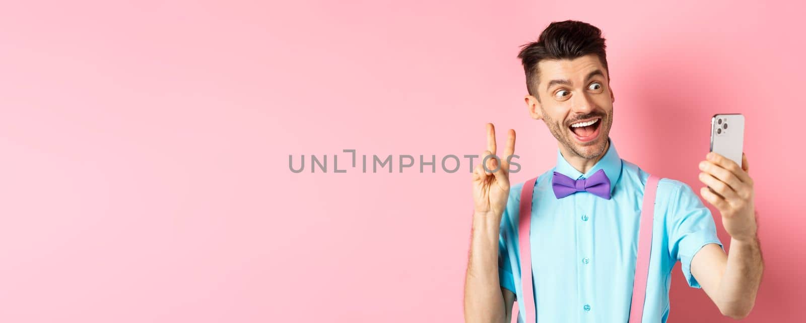 Technology concept. Funny man with moustache and bow-tie taking selfie on smartphone, showing peace sign and smiling at mobile camera, pink background.