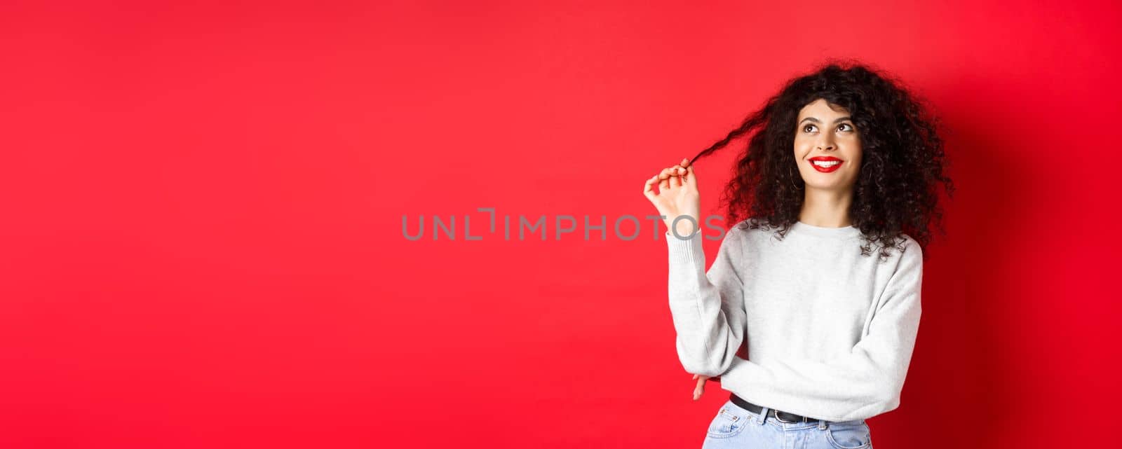 Beautiful female model playing with curly and looking happy up, thinking dreamy about something, standing against red background.