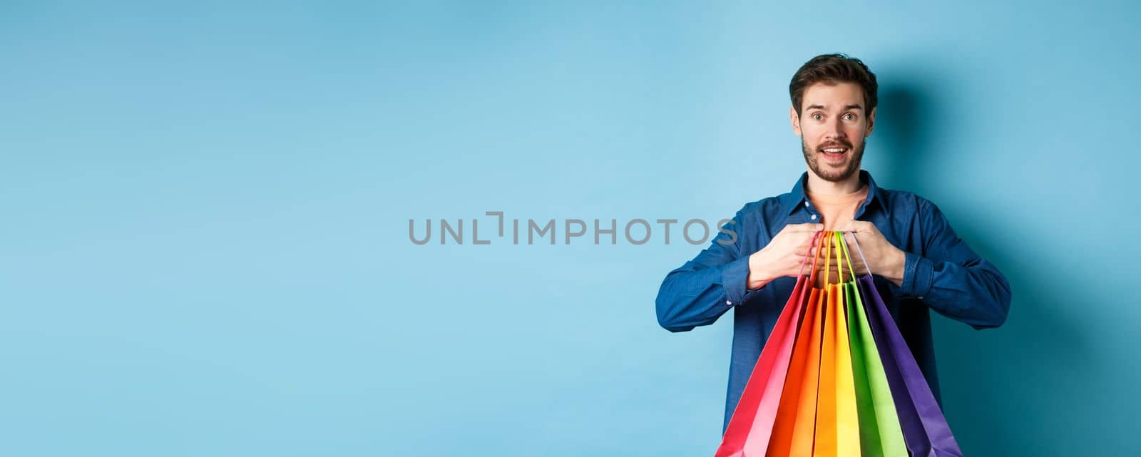 Happy young man holding colorful shopping bags and smiling excited, buying gifts, standing on blue background.