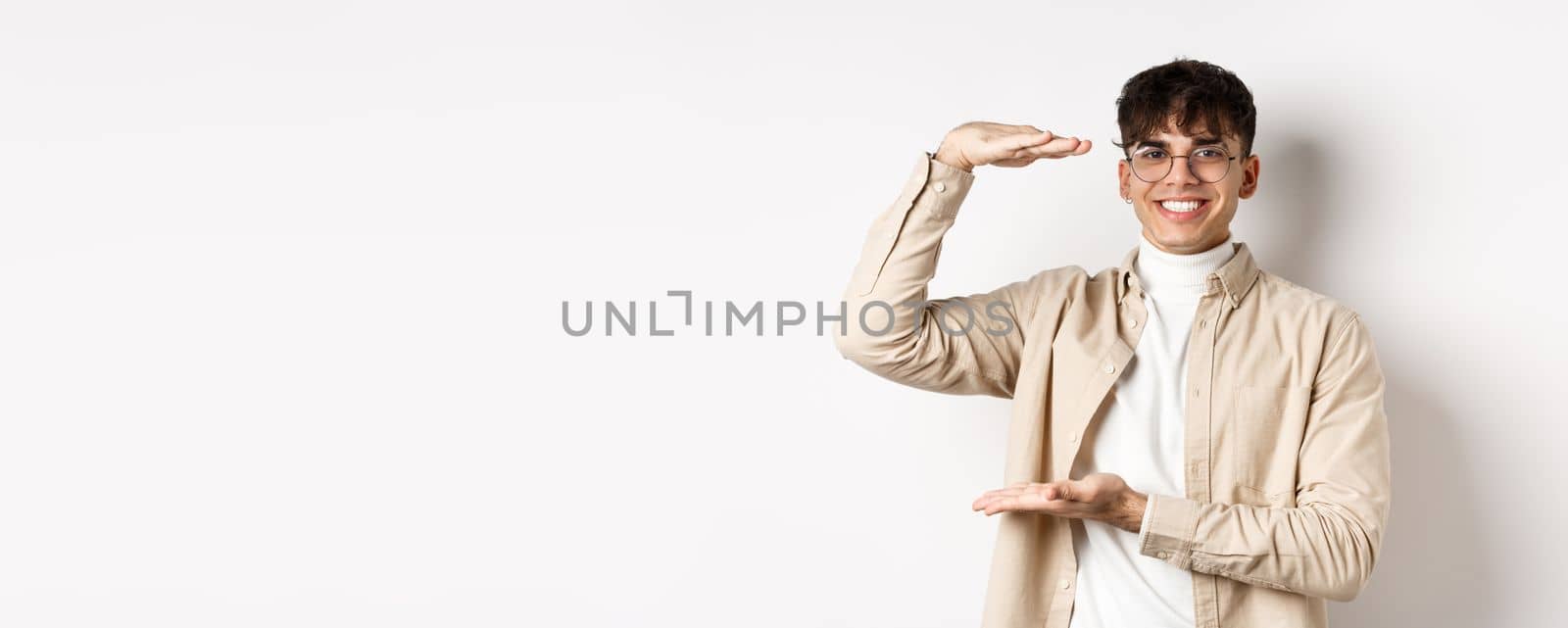 Real people. Happy modern guy in glasses showing big size, shaping box and smiling, showing something large, standing on white background.