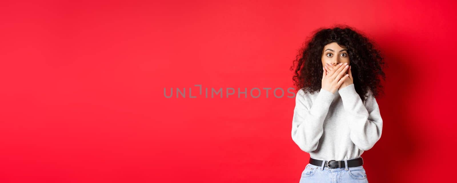 Image of speechless caucasian woman covering mouth with hands, looking shocked at camera, standing in casual clothes on red background.