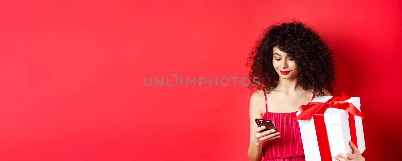 Valentines day and shopping. Beautiful caucasian woman in red dress, holding gift box from lover and using mobile phone, reading message on smartphone, studio background by Benzoix