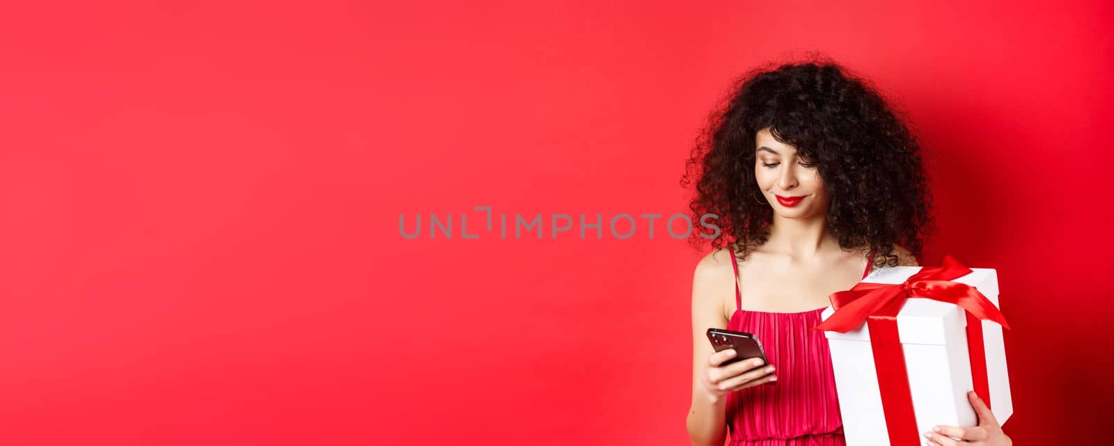 Valentines day and shopping. Beautiful caucasian woman in red dress, holding gift box from lover and using mobile phone, reading message on smartphone, studio background.