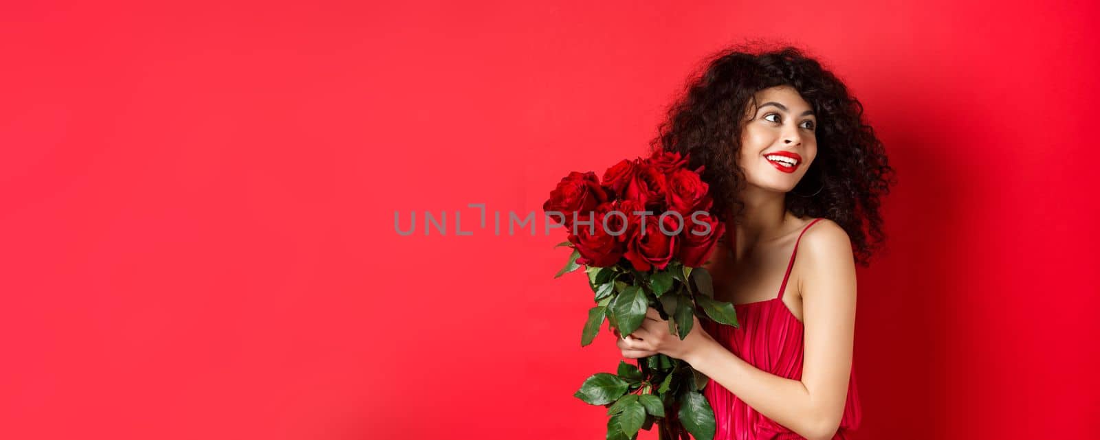 Tender young woman in elegant red dress, holding romantic bouquet of red roses and looking right, smiling dreamy, thinking about lover on Valentines day, studio background.