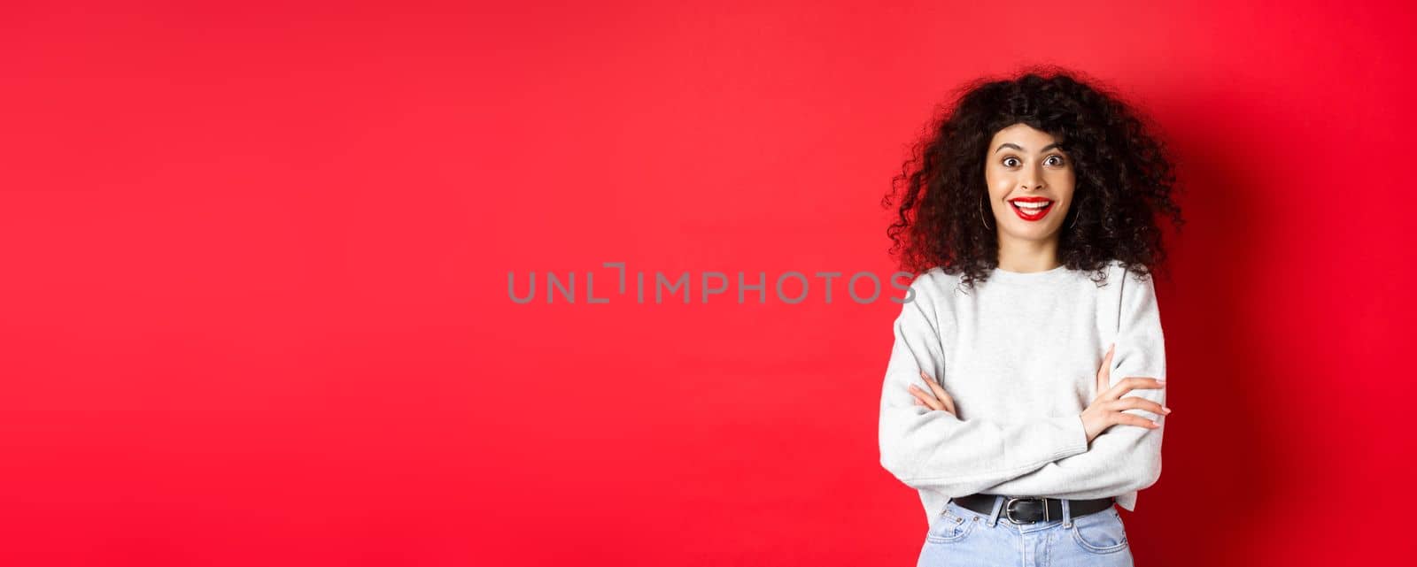 Cheerful young woman with curly hairstyle, raising eyebrows and looking surprised, hear interesting news, red background.