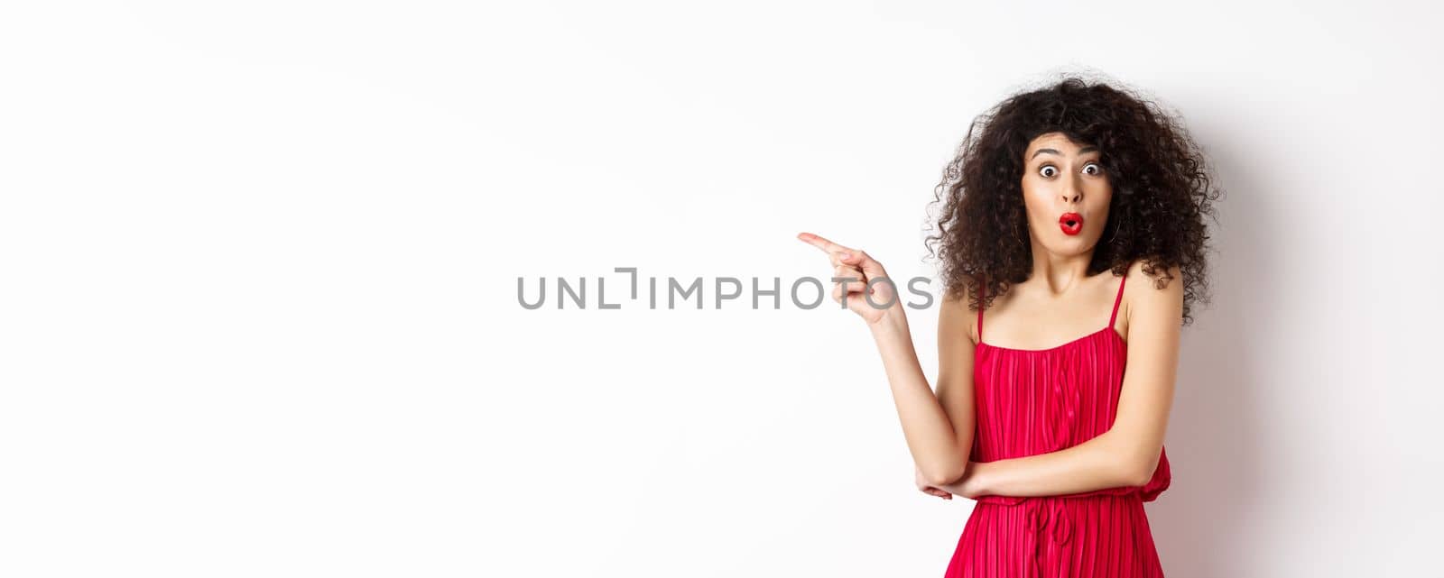 Surprised elegant woman in red dress, saying wow and pointing finger left, showing interesting promo, standing over white background.