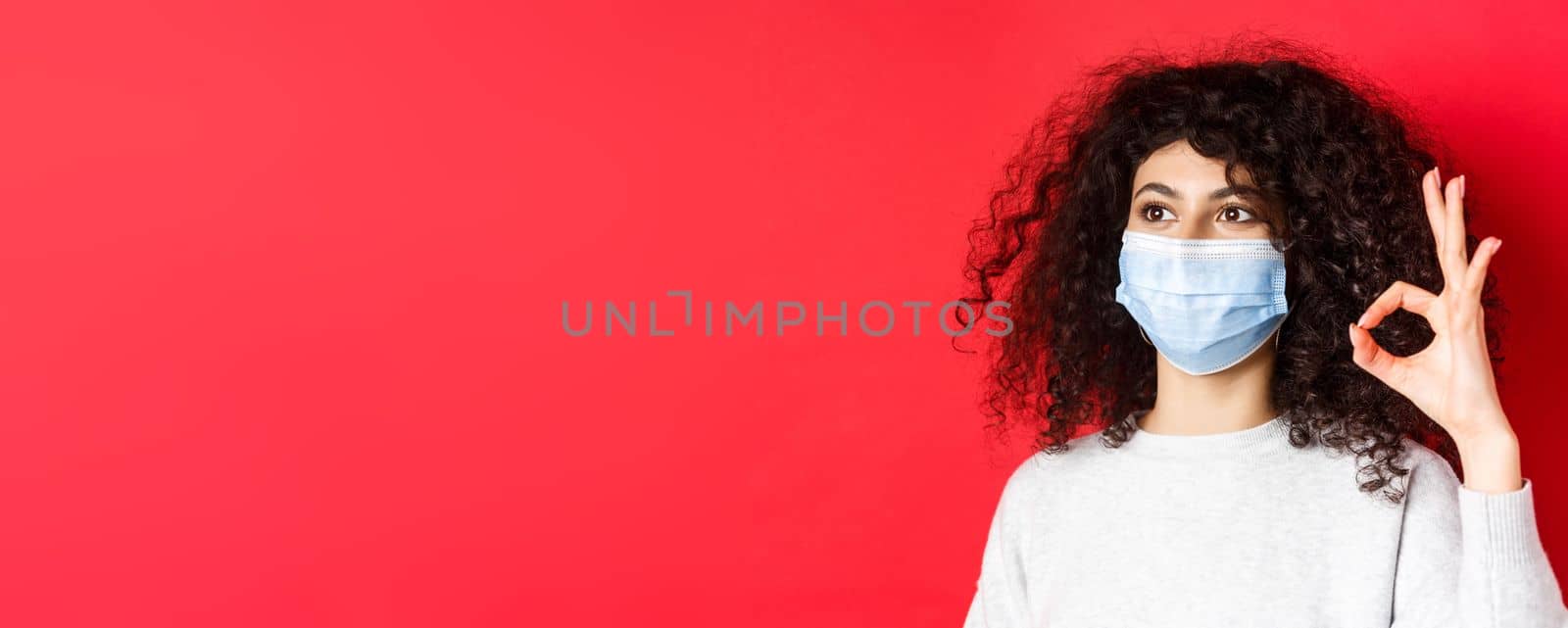 Covid and health concept. Image of young woman in medical mask approve promo, showing okay gesture and looking at empty space, standing on red background.