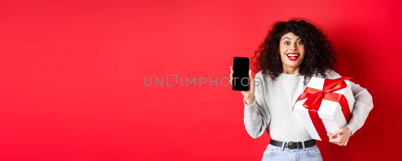 Valentines and lovers day. Excited smiling woman with curly dark hair, showing smartphone empty screen and holding surprise gift on holiday, showing online promo, red background.