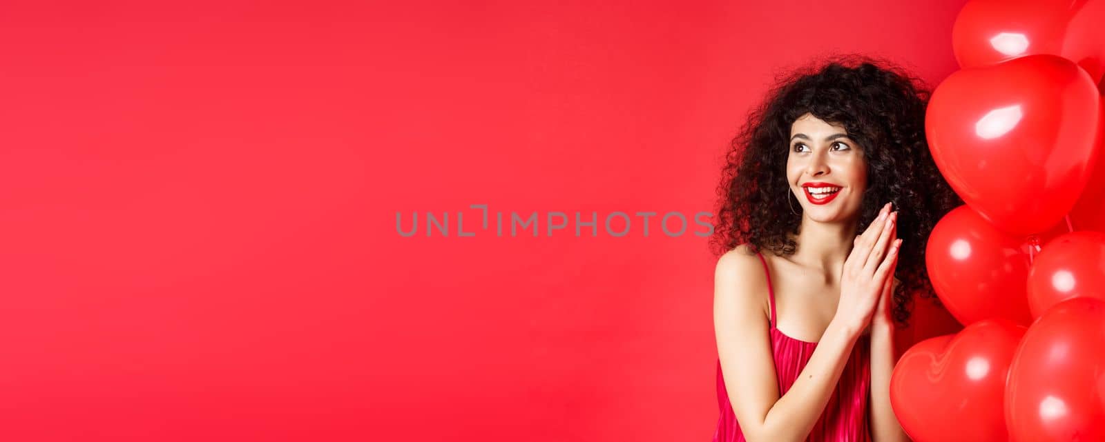 Excited beautiful woman with curly hair, standing near heart balloons and rubbing palms together, expect good deal or relish, red background by Benzoix