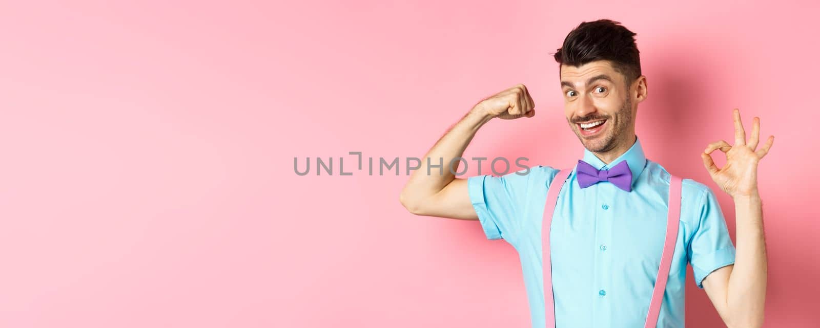 Handsome young man with moustache and bow-tie, flex biceps, showing strong arm and okay sign, workout at gym, standing on pink background.