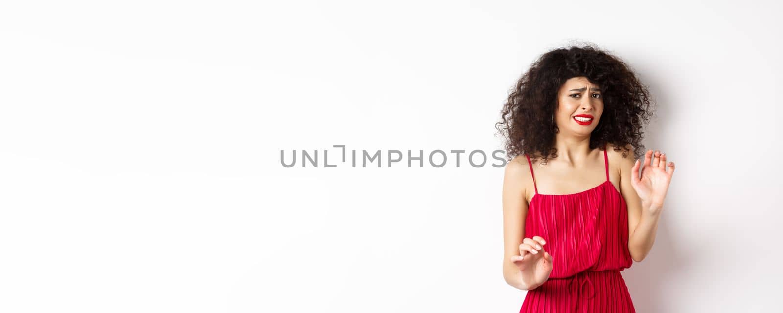 Young woman cringe of something creepy, grimacing and recoil from disgusting awful thing, standing in red dress on white background.