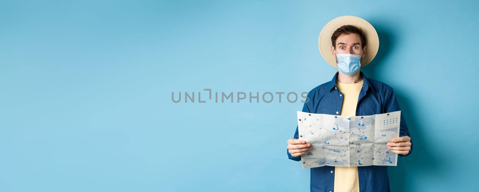 Covid-19, pandemic and travel concept. Tourist in medical mask look surprised, holding map, standing on blue background.