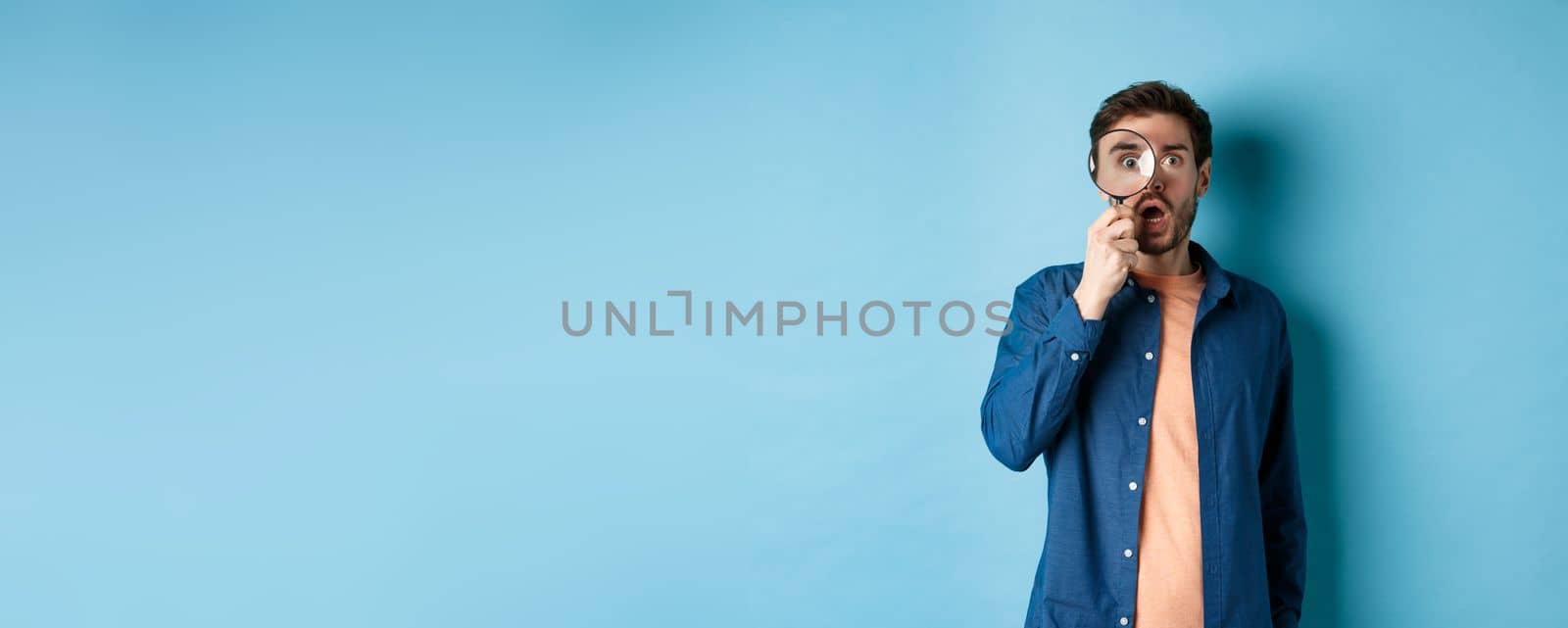 Impressed man look in awe through magnifying glass, drop jaw and stare at camera, standing on blue background.