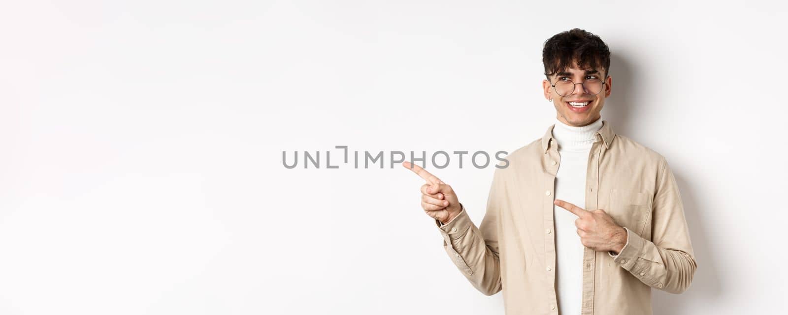 Handsome male model in glasses and trendy clothes, looking and pointing left at empty space, smiling with happy and pleased face expression, white background.