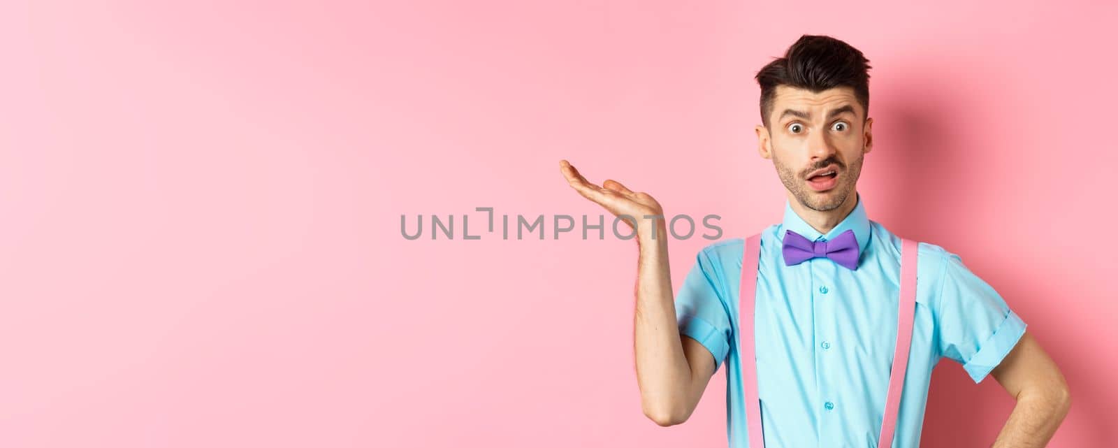 Confused guy cant understand what happening, looking with wtf face, raising hand and staring at camera shocked, standing over pink background.