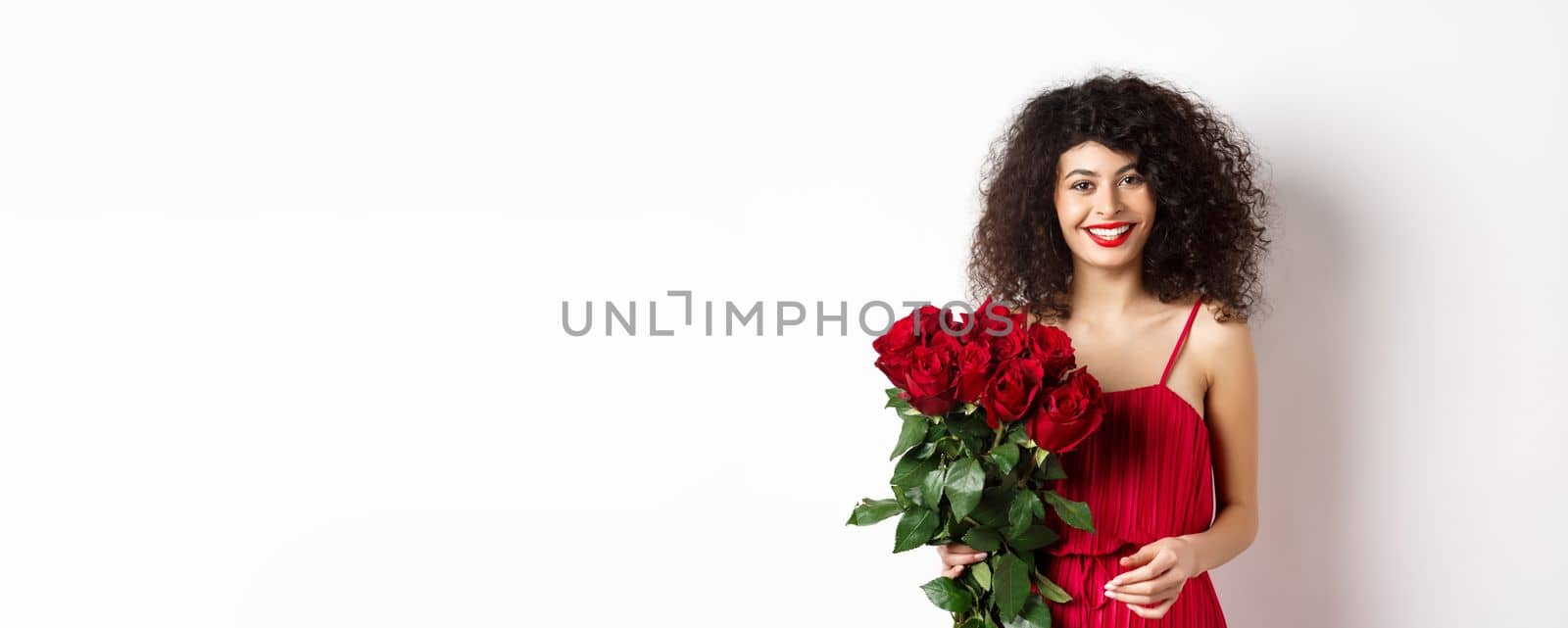 Elegant smiling lady with red lips and dress, holding bouquet of roses and looking happy, white background.