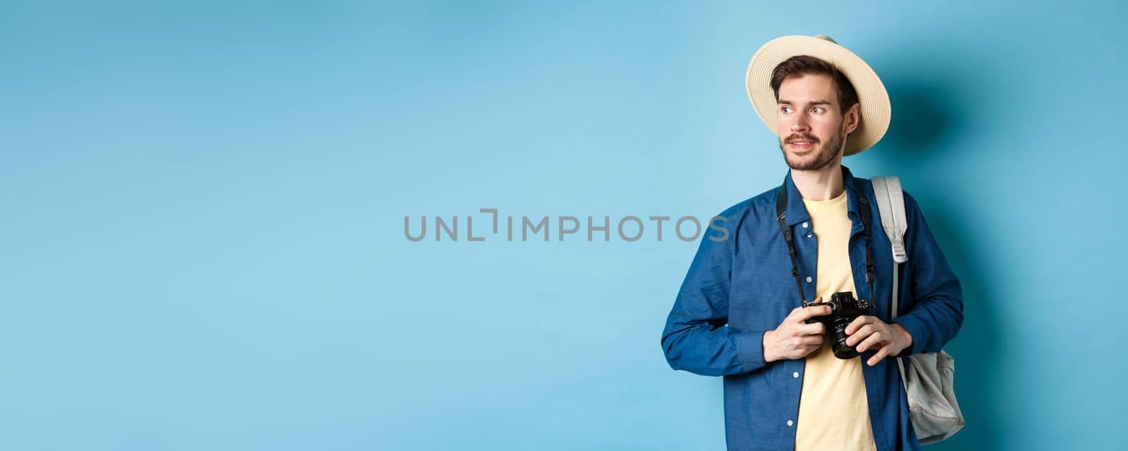 Handsome positive guy in summer hat, holding photo camera and looking aside, tourist taking pictures on vacation, standing with backpack on blue background.