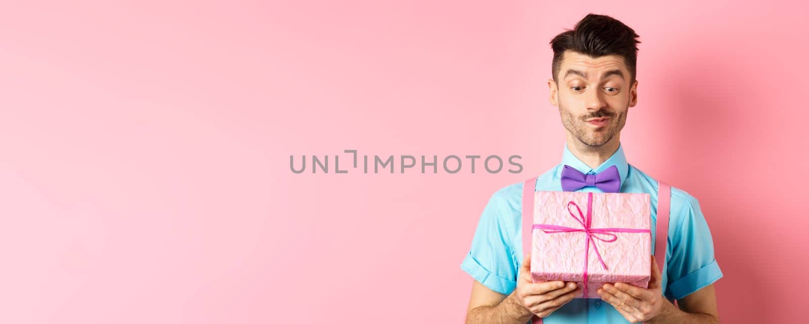 Holidays and celebration concept. Happy smiling guy receive white day gift from his valentine, looking at present with intrigued face, standing over pink background.