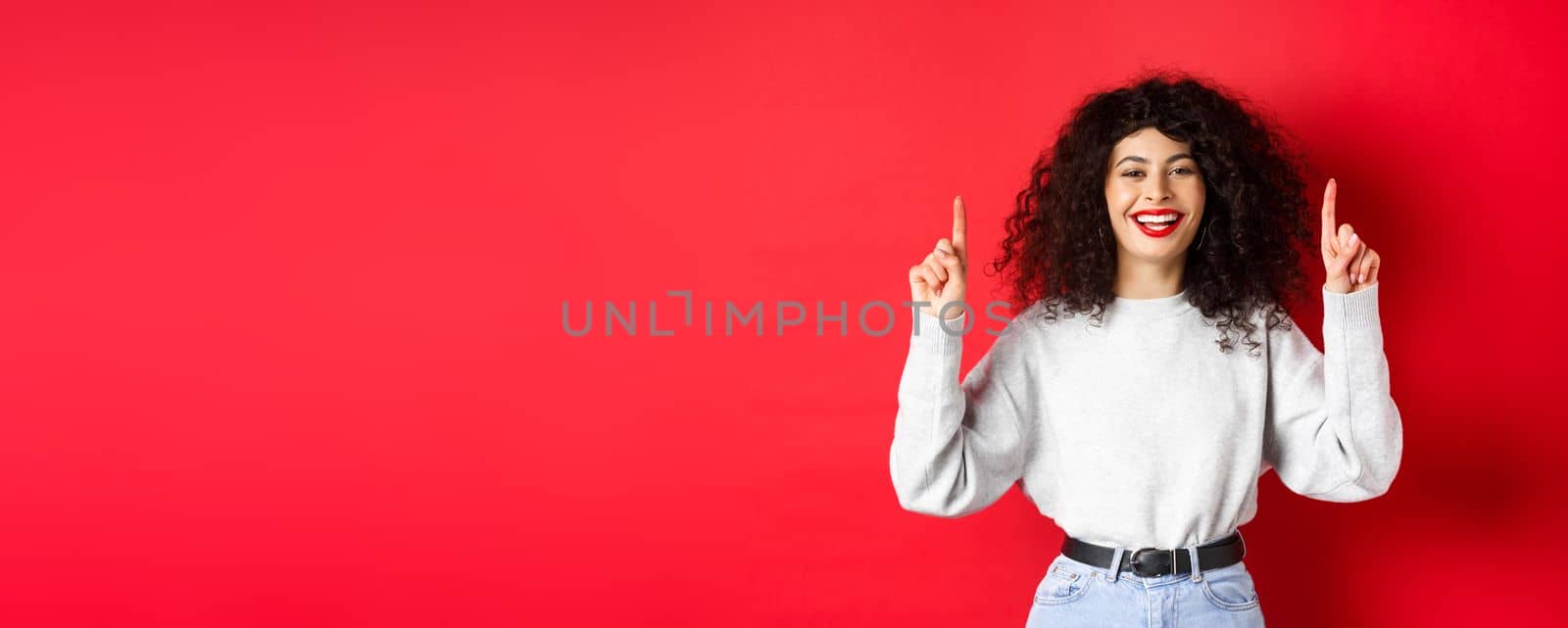 Happy young woman with curly hair, pointing fingers up and laughing, showing perfect white smile, standing against red background.