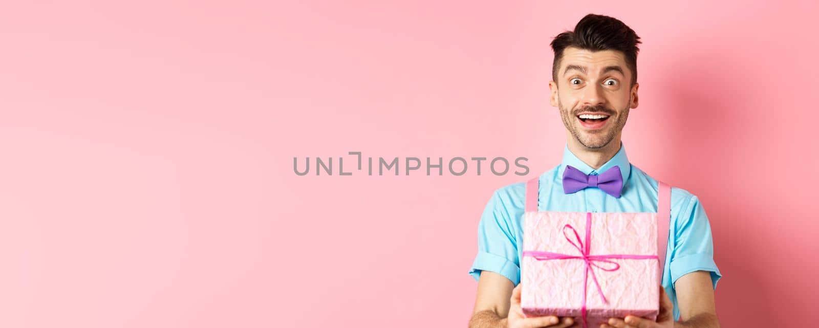 Holidays and celebration concept. Cheerful guy wishing happy birthday and giving you gift wrapped in box, standing over pink background in festive clothes by Benzoix