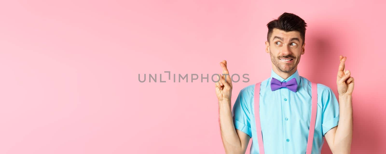 Hopeful handsome guy in bow-tie making wish, cross fingers for good luck and looking left, waiting for results or praying, standing on pink background.