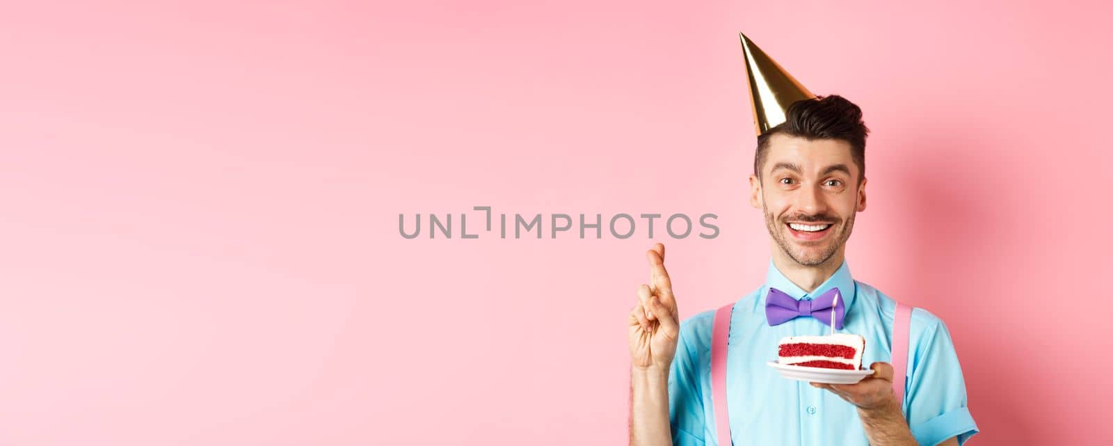 Holidays and celebration concept. Happy young man enjoying birthday party, wearing cone hat and cross fingers, making wish on bday cake with candle, pink background.