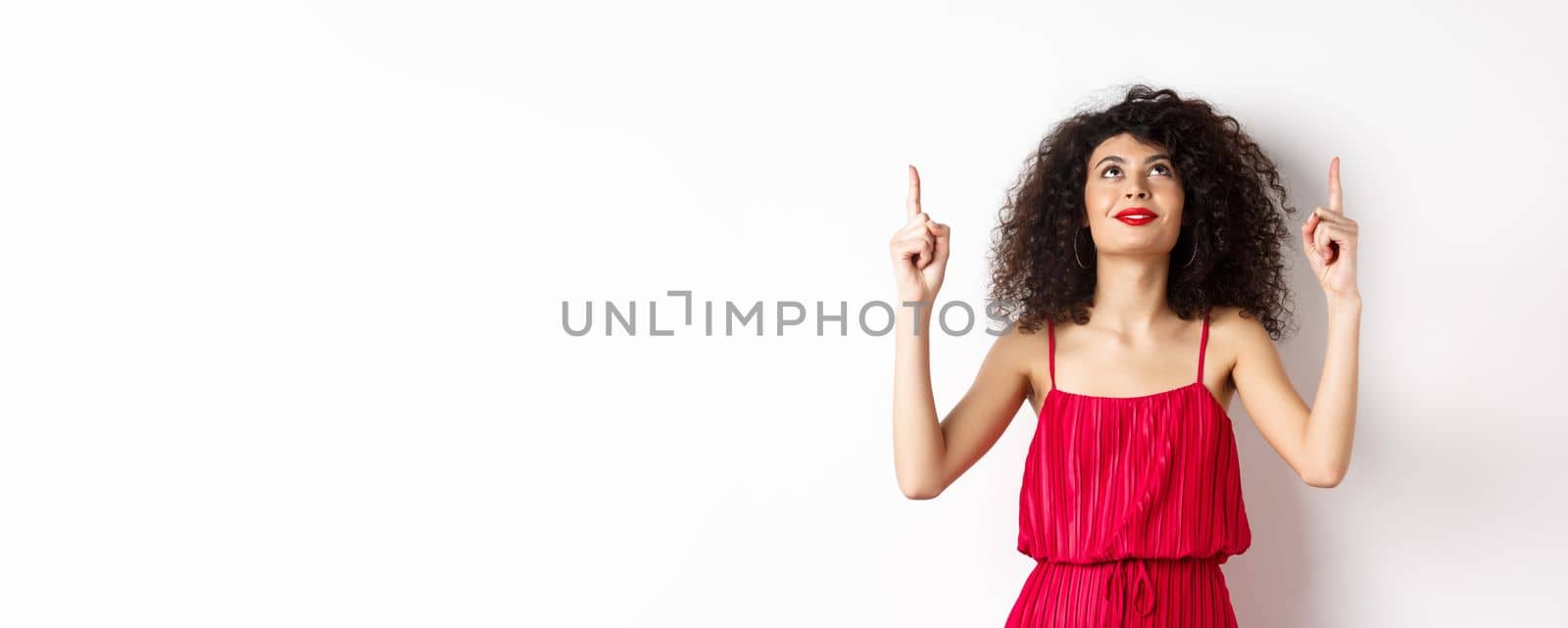 Dreamy young woman in trendy red dress, looking up and pointing at logo, smiling happy, standing over white background. Copy space