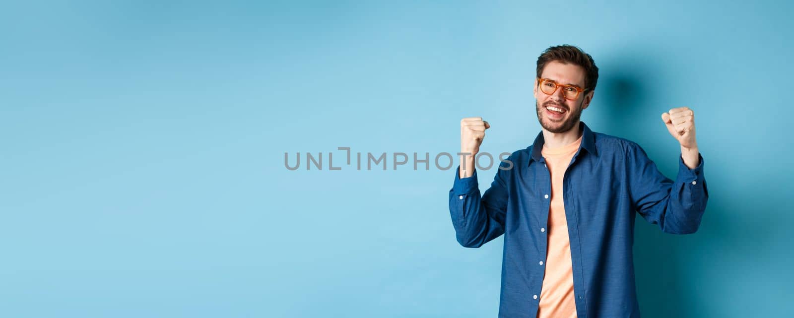 Cheerful young man celebrating, raising hands up and saying yes, wearing eyewear, standing on blue background.