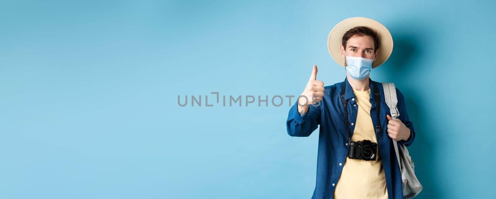 Covid-19 and summer holidays concept. Cheerful tourist in straw hat and medical mask praise and recommend good vacation hotel, showing thumb up, going on travel with backpack by Benzoix