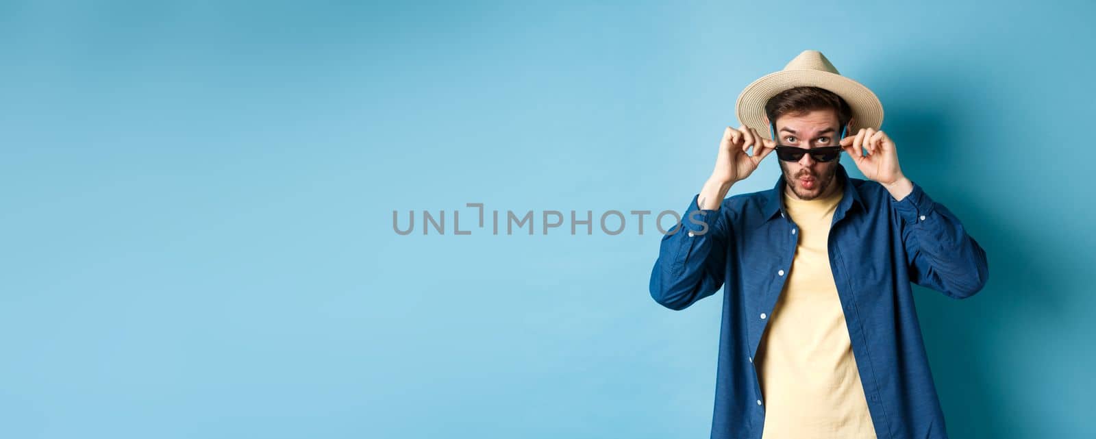 Portrait of tourist takes-off sunglasses and saying wow, staring impressed at camera, checking out cool thing, standing in summer hat on blue background.