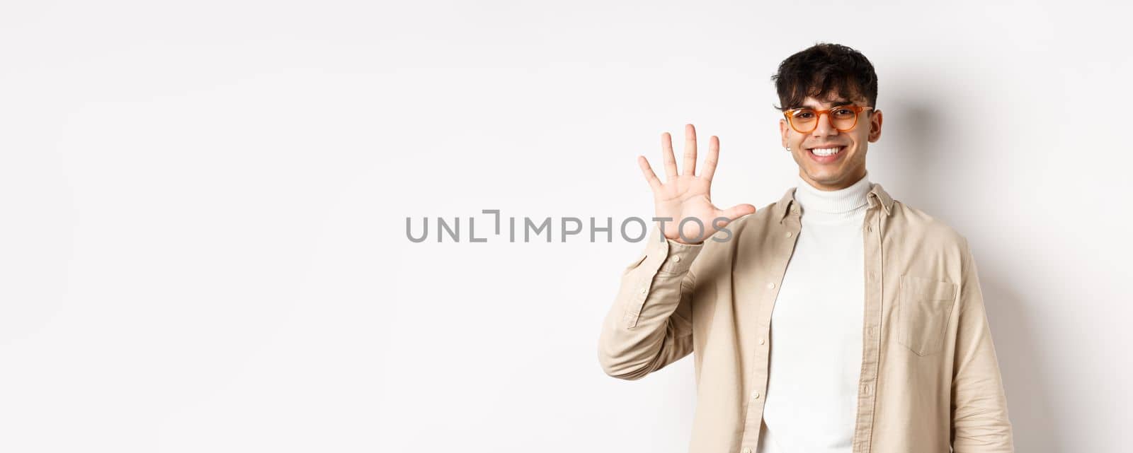 Modern guy in glasses and stylish outfit, showing five fingers number and smiling, making order, standing on white background.