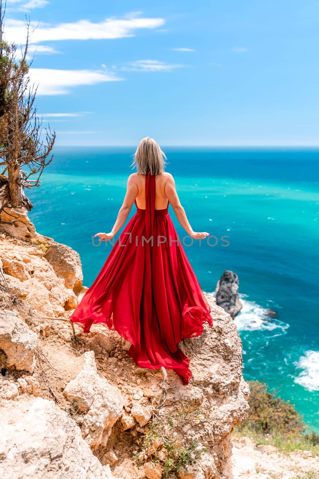 A girl with loose hair in a long red dress descends the stairs between the yellow rocks overlooking the sea. A rock can be seen in the sea. Sunny path on the sea from the rising sun by Matiunina