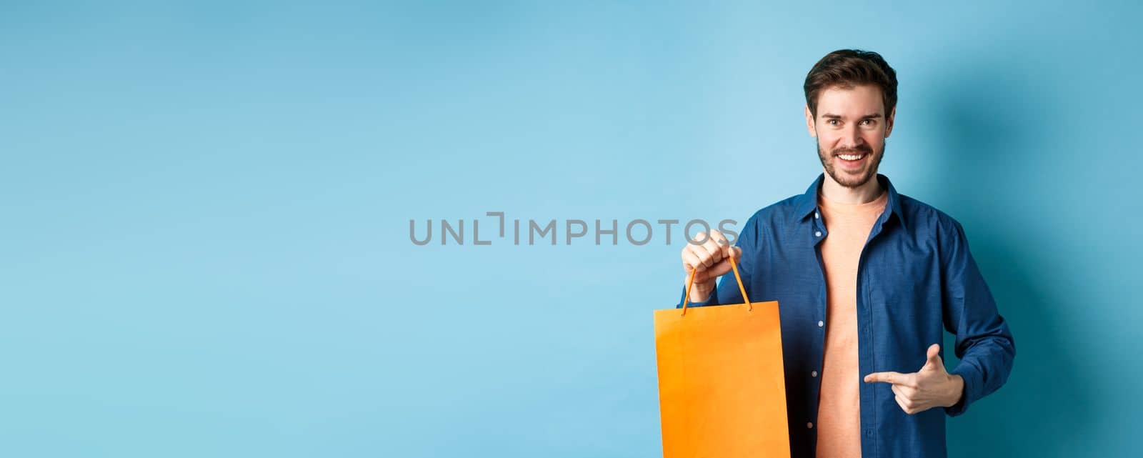 Smiling caucasian man pointing finger at orange shopping bag with bought items, standing on blue background by Benzoix