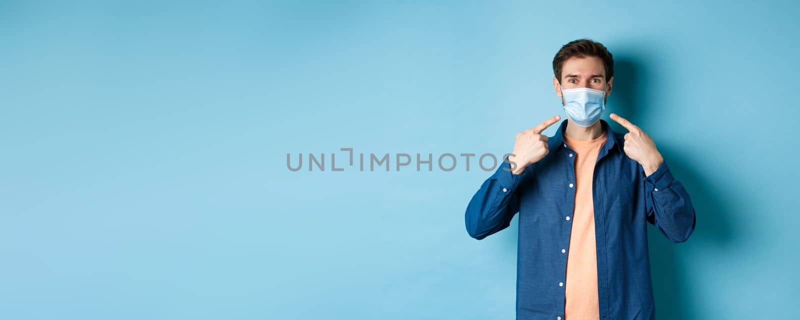 Coronavirus and pandemic concept. Portrait of happy and healthy man pointing fingers at his medical mask, smiling at camera, standing on blue background by Benzoix