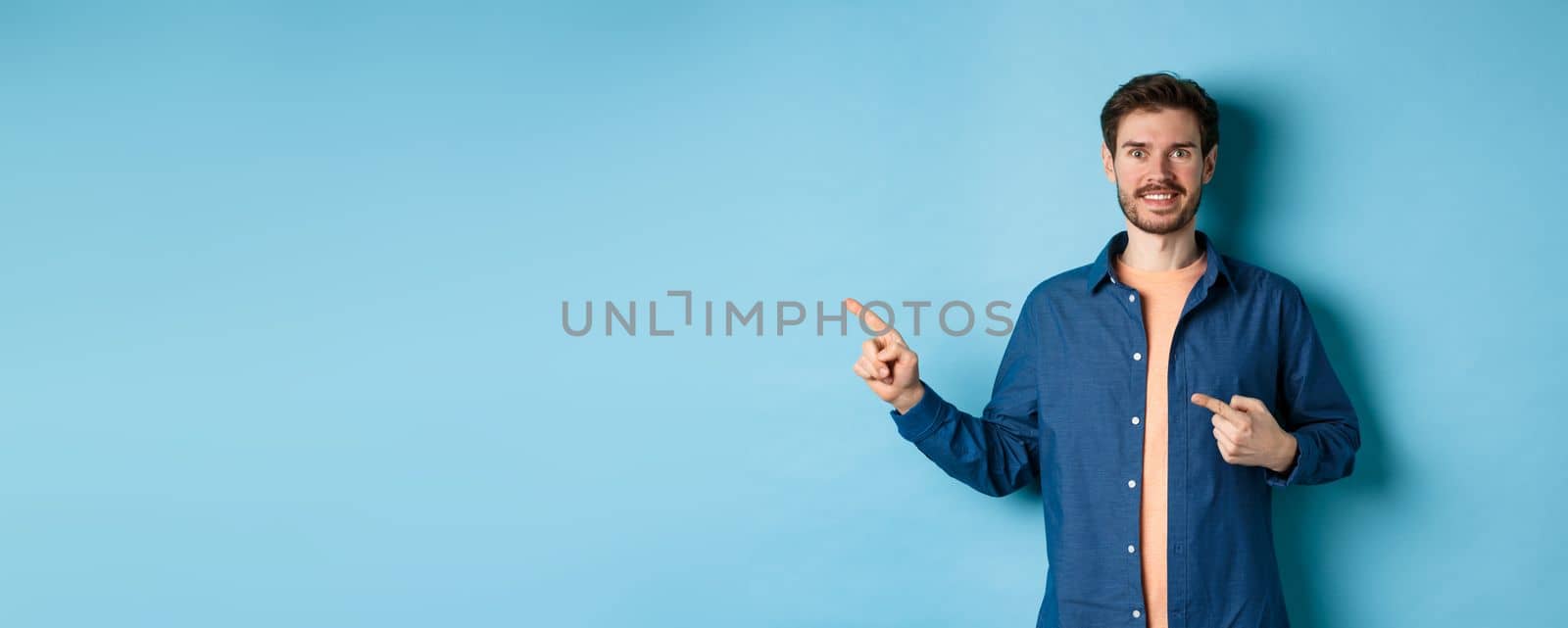 Happy smiling caucasian man showing advertisement, pointing fingers left at empty space, standing on blue background.