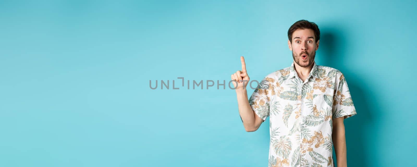 Surprised caucasian tourist in hawaiian shirt, pointing finger up and say wow, checking out promotion, standing on blue background.