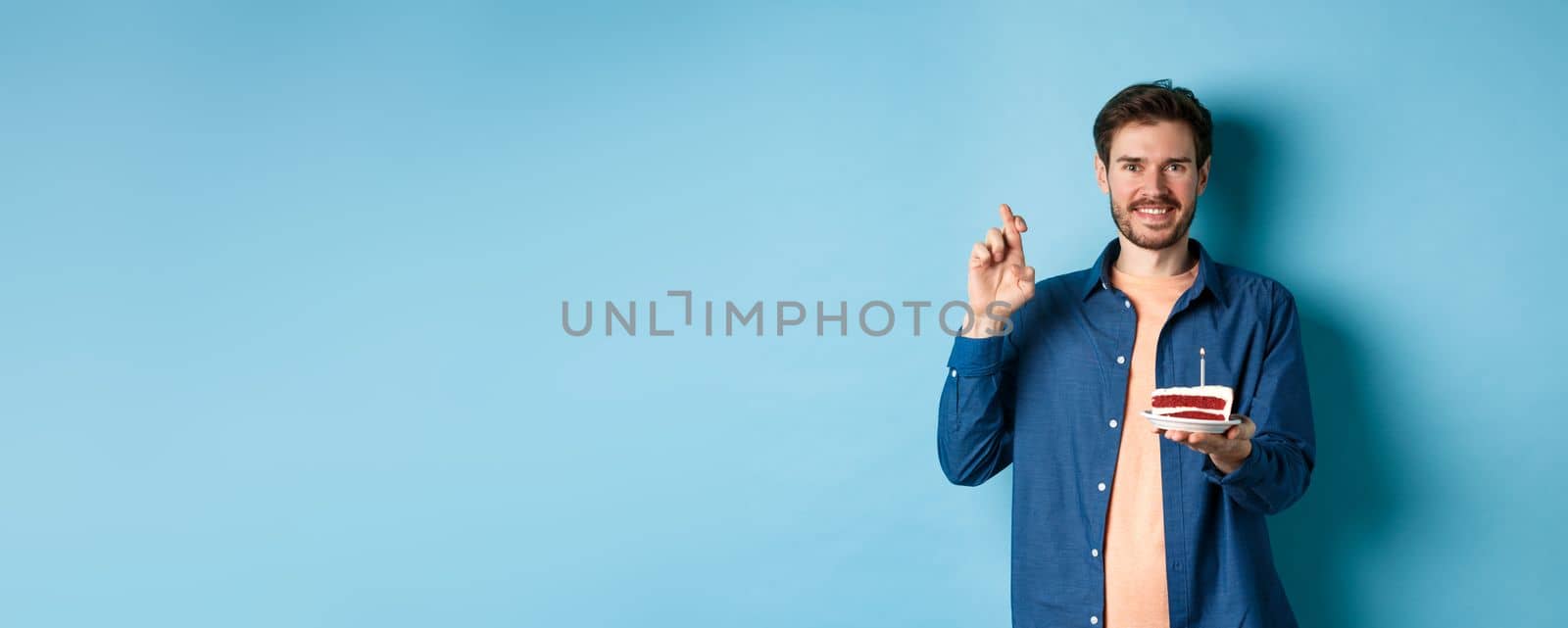 Celebration and holiday concept. Hopeful young man making wish on birthday cake, cross fingers for good luck and smiling positive, standing on blue background by Benzoix