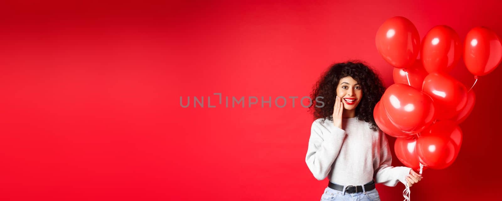 Holidays and celebration. Surprised birthday girl holding helium party balloons and looking touched at camera, being congratulated, red background by Benzoix