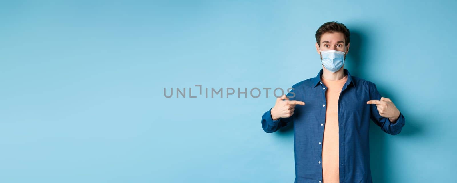 Covid-19 and pandemic concept. Healthy young man in medical mask pointing fingers at center logo, standing on blue background by Benzoix